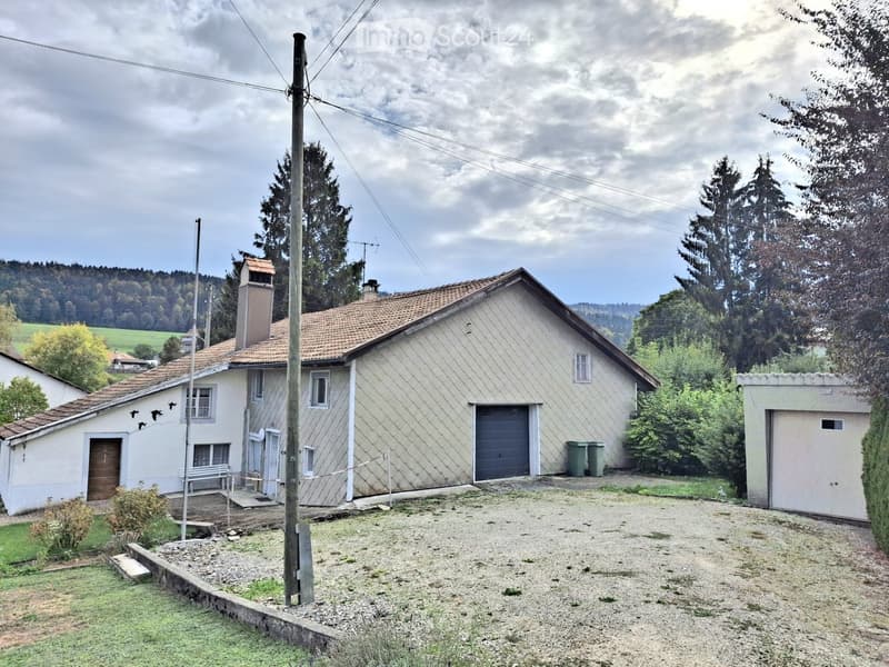 Two-story house with beige siding, red-tiled roof, garage, and small outbuilding. The house has multiple windows and a chimney, surrounded by trees and a gravel driveway.