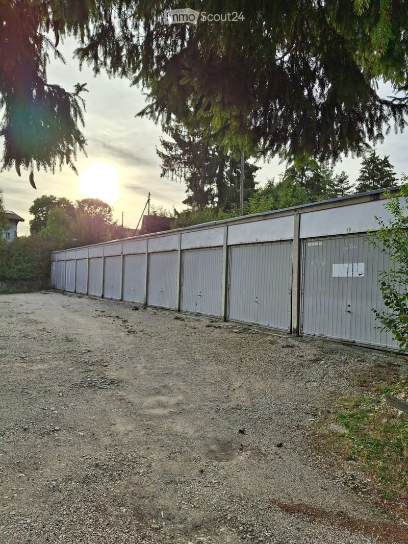 A row of grey storage units with closed doors, situated on a gravel surface surrounded by trees and greenery.