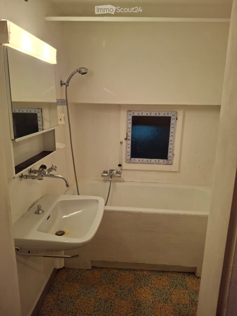 Bathroom with a sink, bathtub, mirror, and showerhead. The walls are tiled, and there is a decorative window above the bathtub.