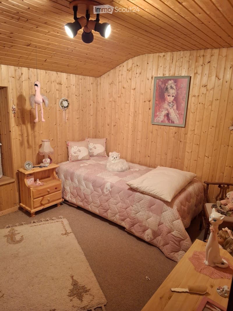 Cozy bedroom with wooden walls and ceiling, featuring a bed with pink bedding, a nightstand with a lamp, a small table with a cat figurine, and a fluffy cat on the bed.