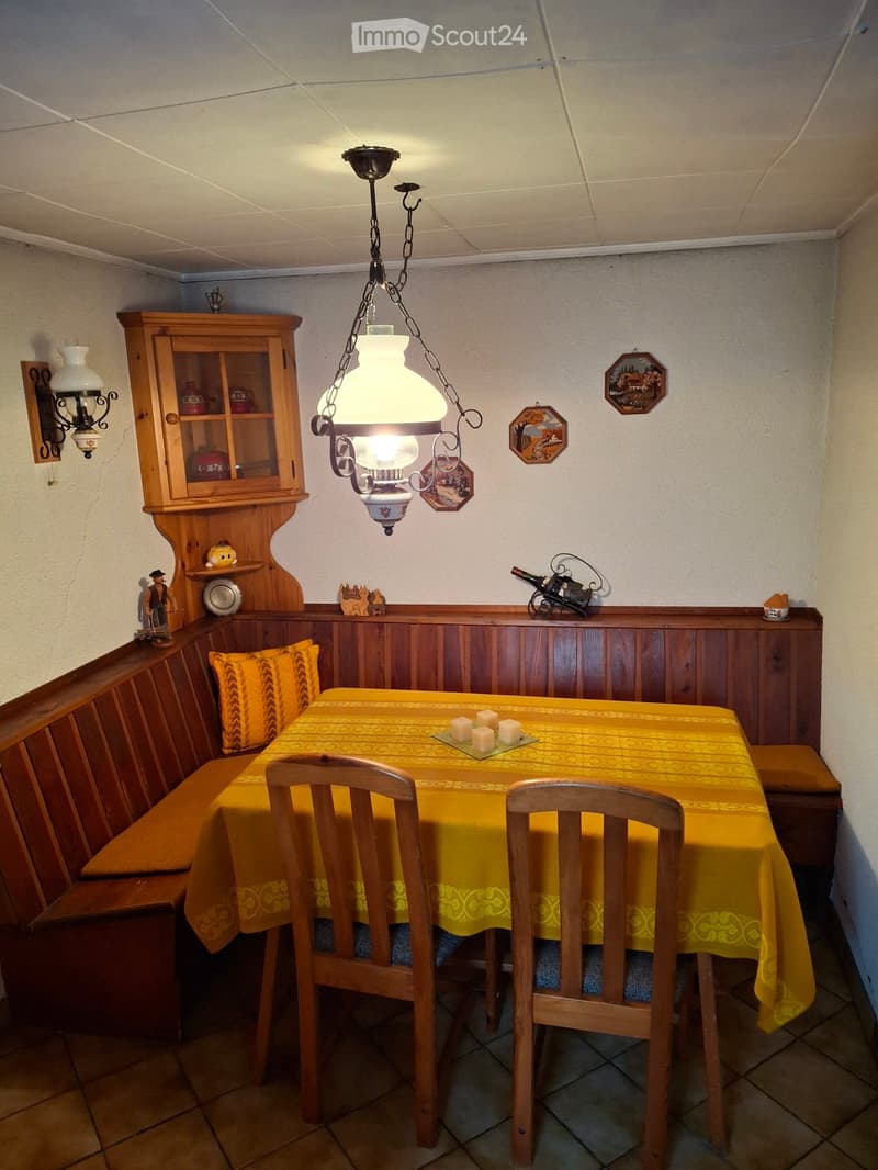 Cozy dining area with a wooden table covered in a yellow tablecloth, two wooden chairs, a built-in wooden bench, a hanging chandelier, a wooden cabinet with glass doors, and decorative items on the walls and shelves.