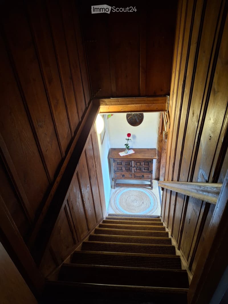 Wooden staircase leading up with wooden railings, a small wooden table with a vase of red rose, a round woven rug, and a wall-mounted clock visible at the top of the stairs.