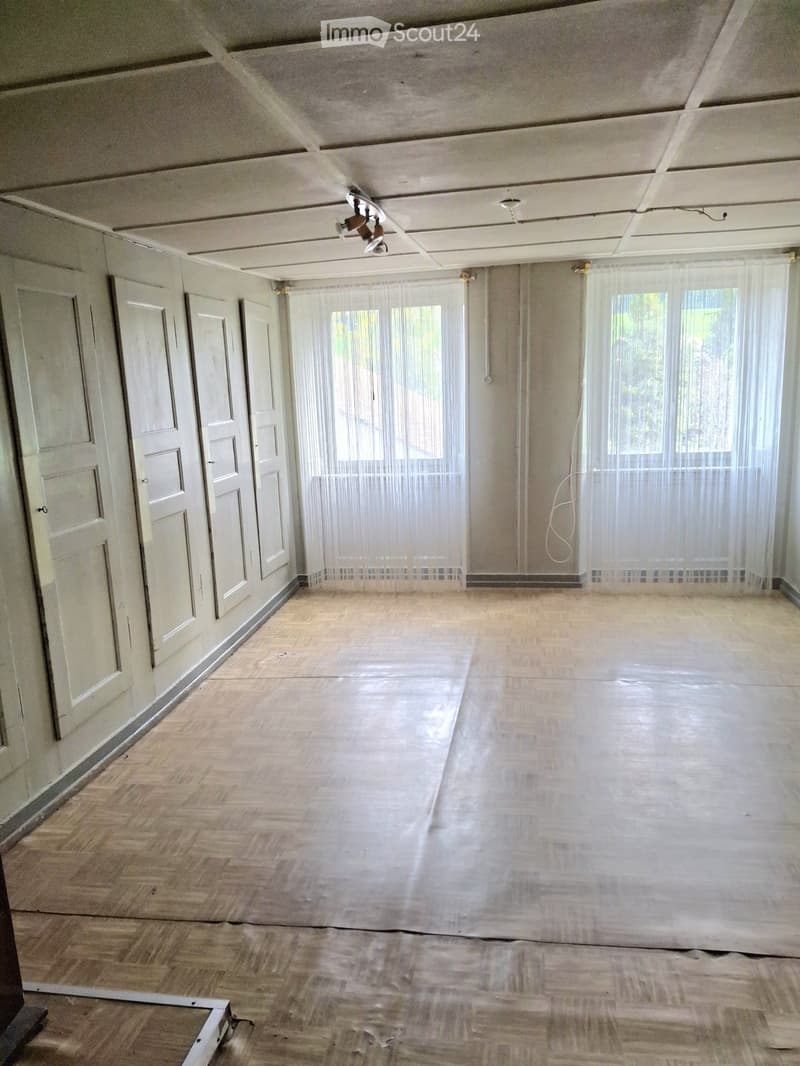 Empty room with wooden flooring, white walls, multiple doors, and large windows covered with sheer curtains.