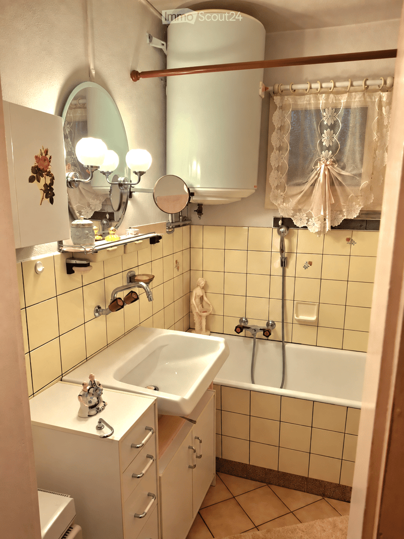 Bathroom with a sink, bathtub, mirror, and cabinet. Features a decorative statue on the sink counter and a window with lace curtains.