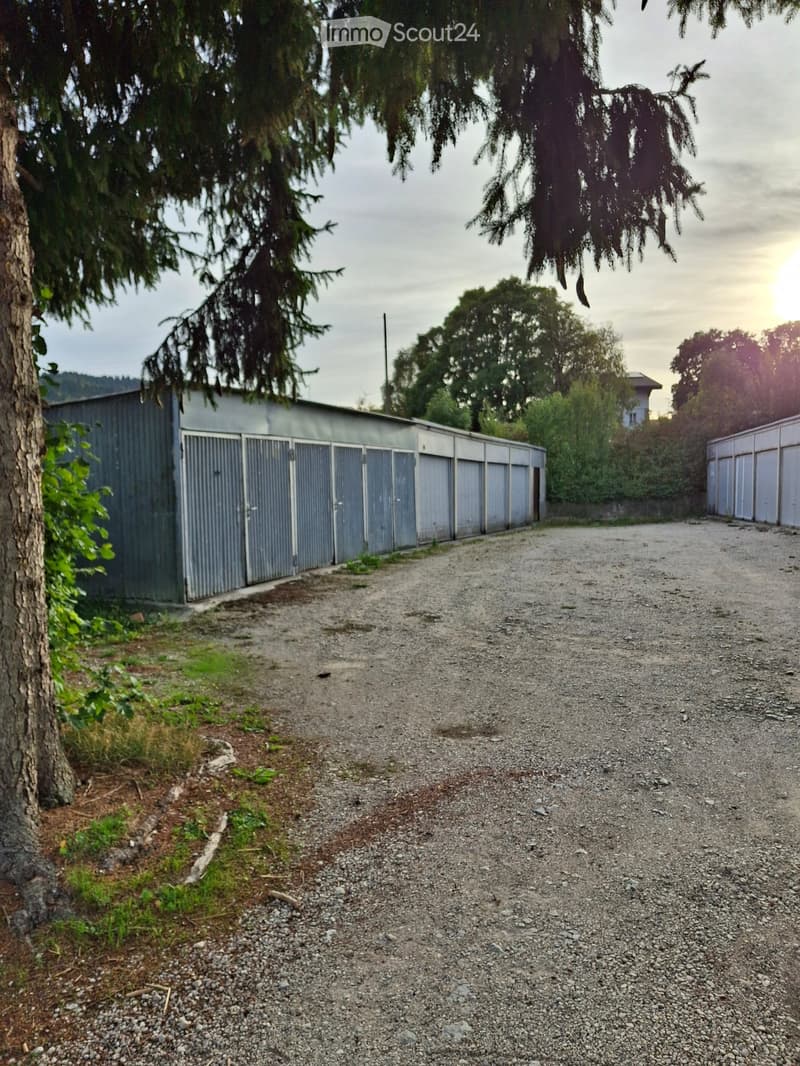 Gravel driveway leading to a row of metal storage units, surrounded by trees and greenery.