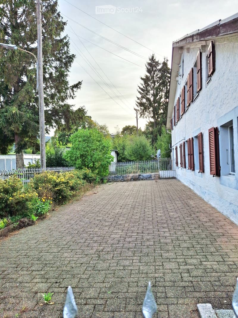 Brick-paved pathway leading to a two-story house with light blue walls and brown shutters, surrounded by greenery and a white fence.