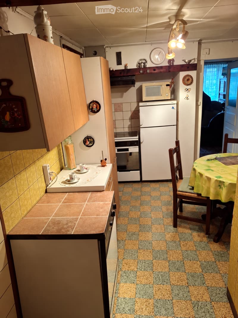 A compact kitchen with a refrigerator, microwave, oven, and a small dining area with a table and chairs. The kitchen features wooden cabinets, a tiled backsplash, and a checkered floor.