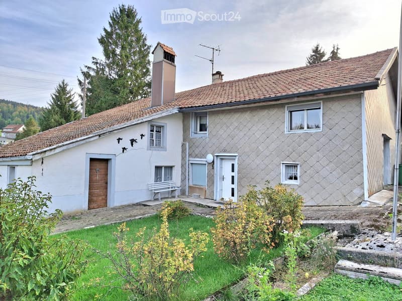 Two-story house with a tiled roof, white exterior walls, multiple windows, and a small garden area with a bench.