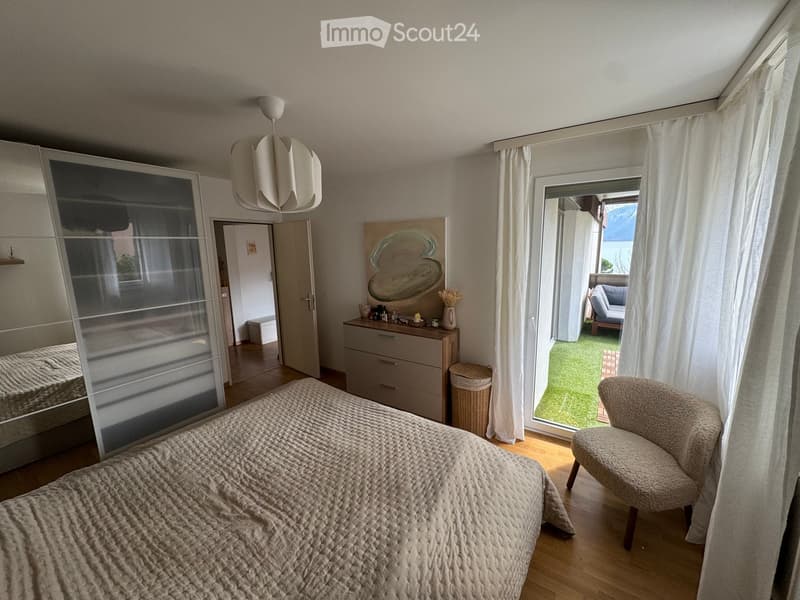 Modern bedroom with a large bed, a plush armchair, a dresser with drawers, a mirror, a hanging lamp, a painting, and a vase on the dresser. The room has wooden flooring and large windows with white curtains, leading to a balcony with a view.