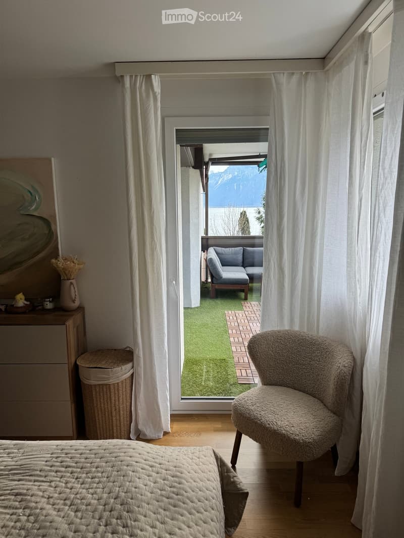 A cozy bedroom with a large bed, a round chair, a curtained window opening to a balcony with a view, and decorative items on a side table.