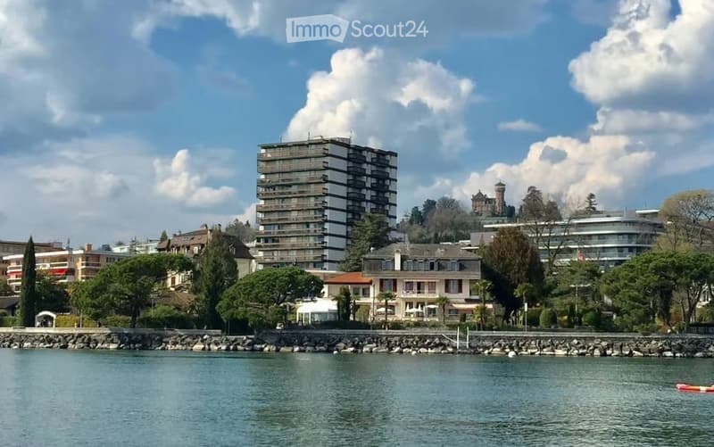 A scenic view of a waterfront area with a mix of residential and commercial buildings, including a prominent high-rise apartment building and a historic castle-like structure in the background, surrounded by trees and a calm lake.