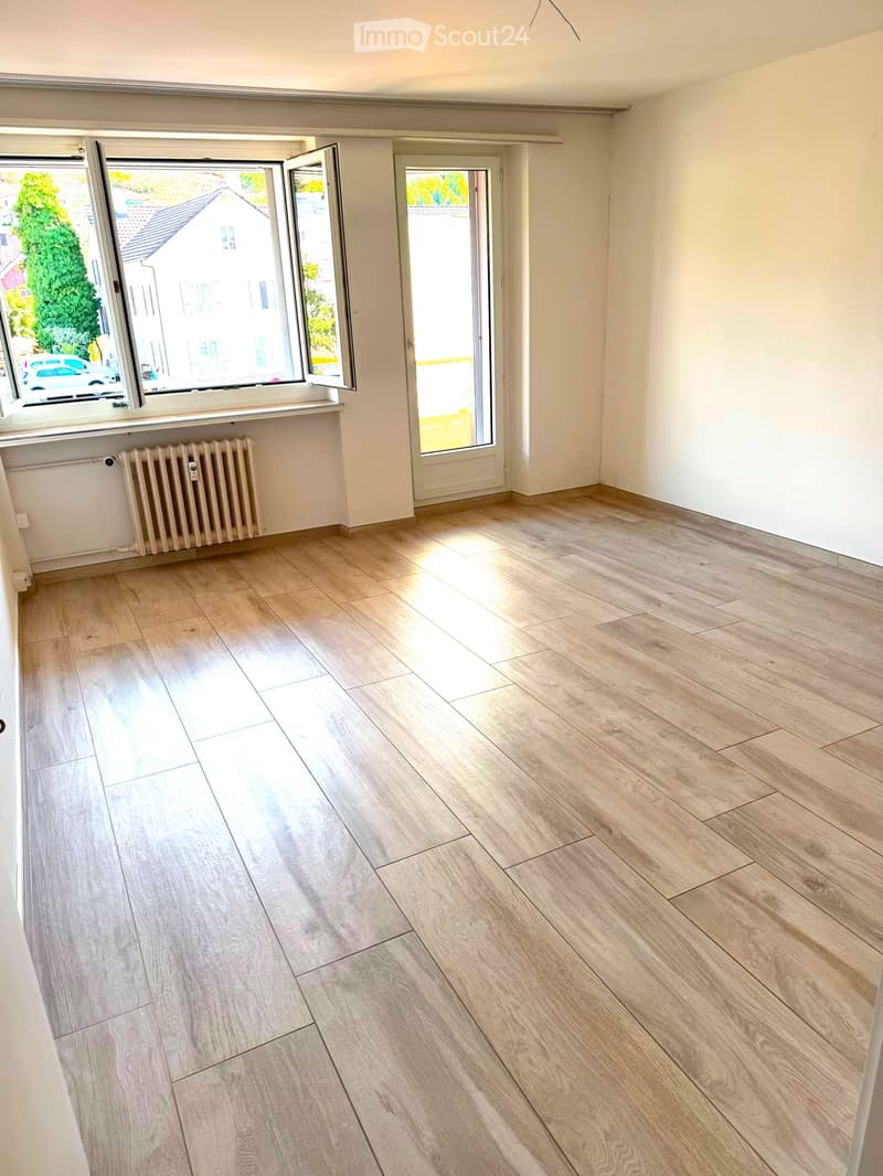 Empty room with light wooden flooring, white walls, large windows, and a radiator. No furniture or equipment present.
