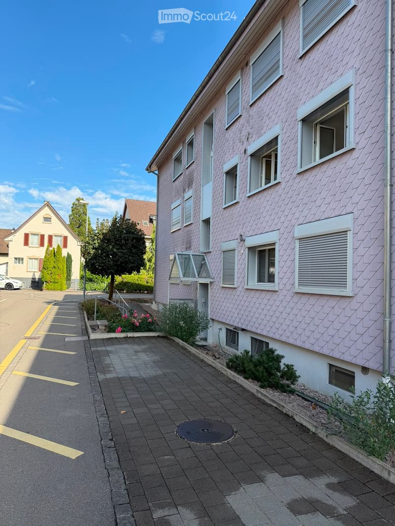 A two-story residential building with pink siding, multiple windows, and a small garden area in front.