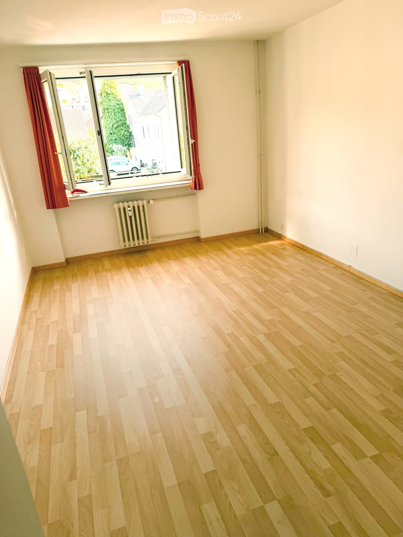Empty room with light wooden flooring, white walls, and a large window with red curtains. A radiator is visible below the window.