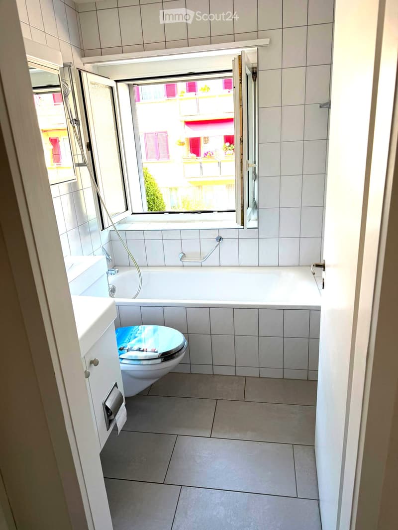 Modern bathroom with white tiled walls and floor, featuring a bathtub, toilet, sink, and a large window with an open view of the street.