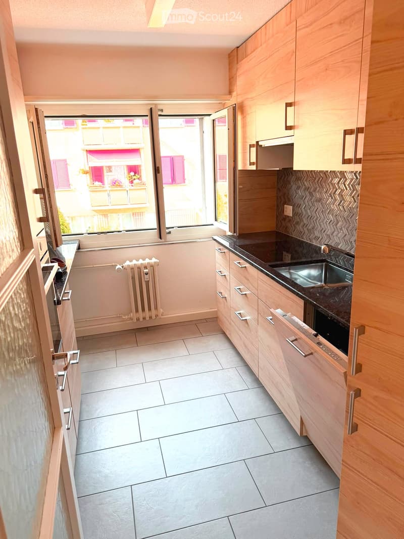 Modern kitchen with light wooden cabinets, dark countertops, and a sink. The space features tiled flooring and a radiator, with a window offering a view of the street.