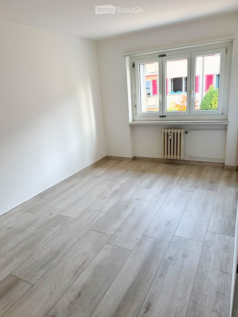 Empty room with light wooden flooring, white walls, and a large window with white frames and a radiator below it.