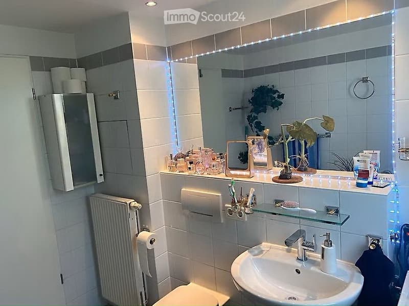 Modern bathroom with white tiles, a large mirror with LED lighting, a sink with a modern faucet, a toilet, and a towel rack. The vanity features a plant and various toiletries.