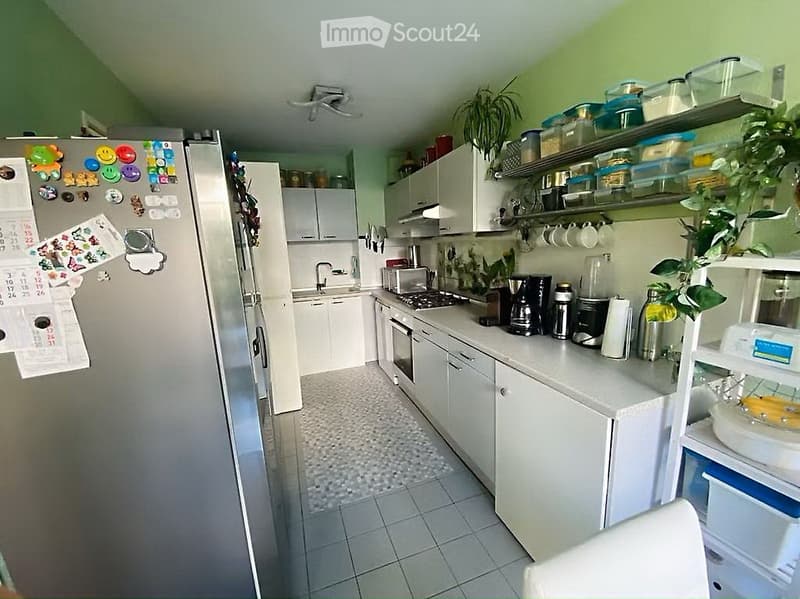 Modern kitchen with white cabinets, stainless steel appliances, and a tiled floor. Features include a refrigerator with magnets, a coffee maker, microwave, and shelves with various items.
