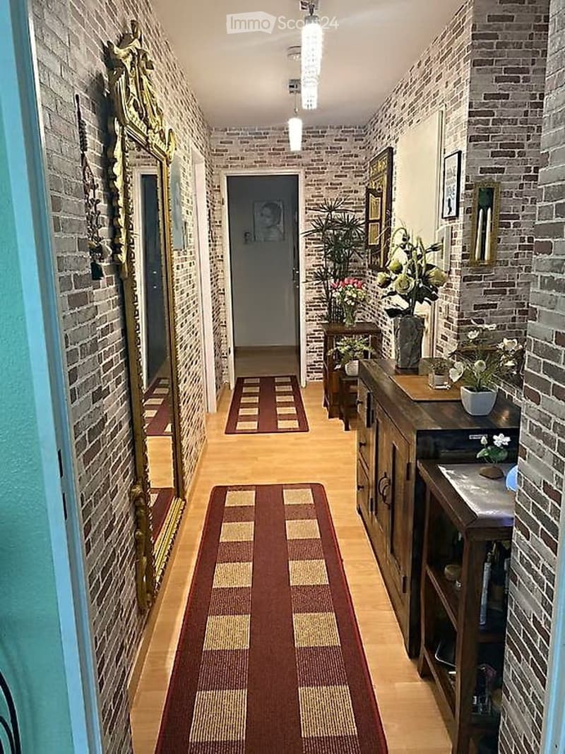 A narrow hallway with brick walls, wooden flooring, and patterned rugs. There are decorative mirrors, framed artwork, and a small console table with potted plants.
