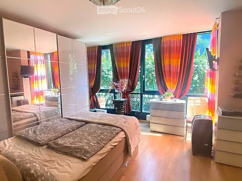Spacious bedroom with a large bed, mirrored wardrobe, nightstand, chair, and large window with colorful curtains.