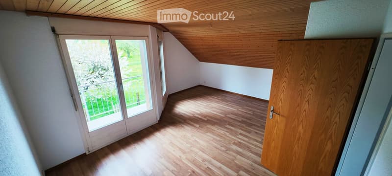 Empty room with wooden flooring, white walls, and a large window with a view of a green outdoor area. The ceiling is slanted with wooden paneling.
