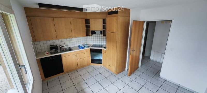 Modern kitchen with light wooden cabinets, tiled backsplash, and stainless steel appliances including a sink, oven, and dishwasher. The space features tiled flooring and an open doorway leading to another room.