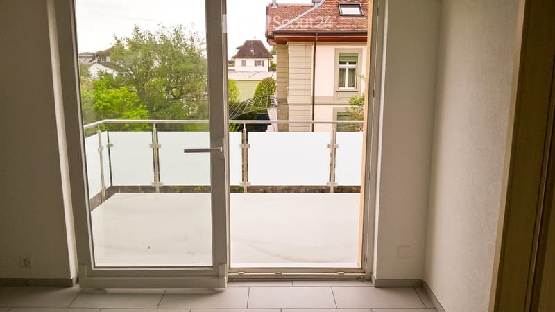 View of a modern balcony with glass sliding doors, white floor, and metal railing, overlooking a residential area with trees and buildings.