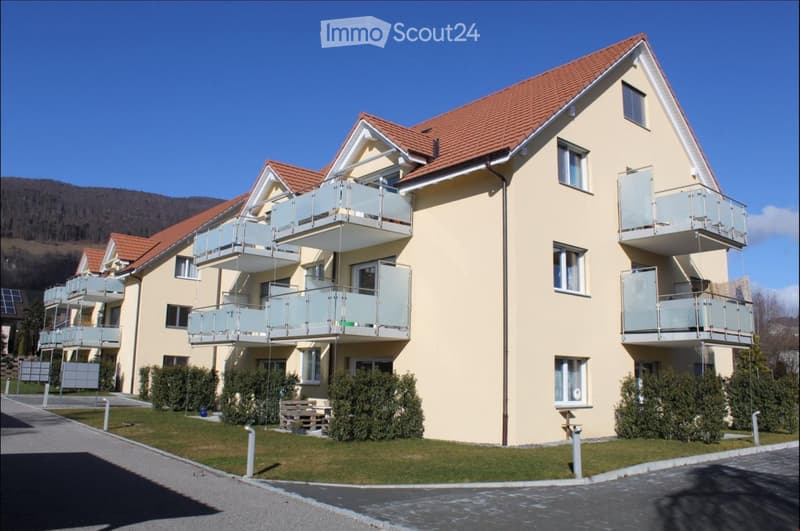 A modern two-story residential building with a red-tiled roof, light yellow walls, and multiple balconies with glass railings. The building is surrounded by neatly trimmed bushes and a paved driveway.