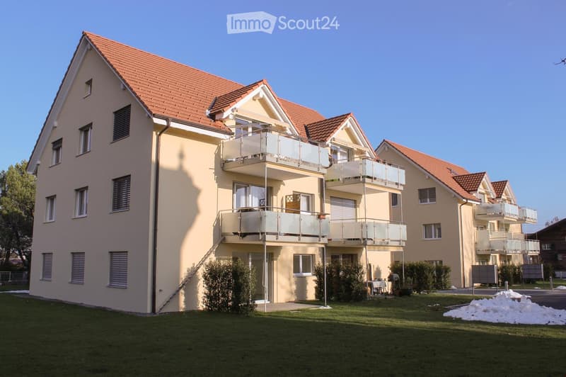 Two-story residential building with beige walls, red-tiled roof, multiple windows, and balconies with glass railings.