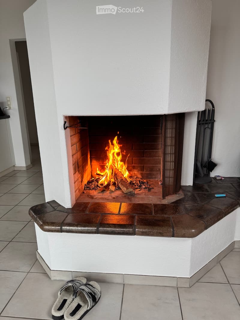 A white brick fireplace with a burning fire, surrounded by a tiled floor and a curved stone hearth. Firewood and fireplace tools are visible, along with a pair of slippers on the floor.