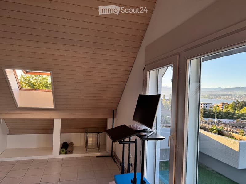 Modern, well-lit room with wooden walls and a skylight, featuring a desk with a large screen, a small table, and yoga mats stored in a niche.