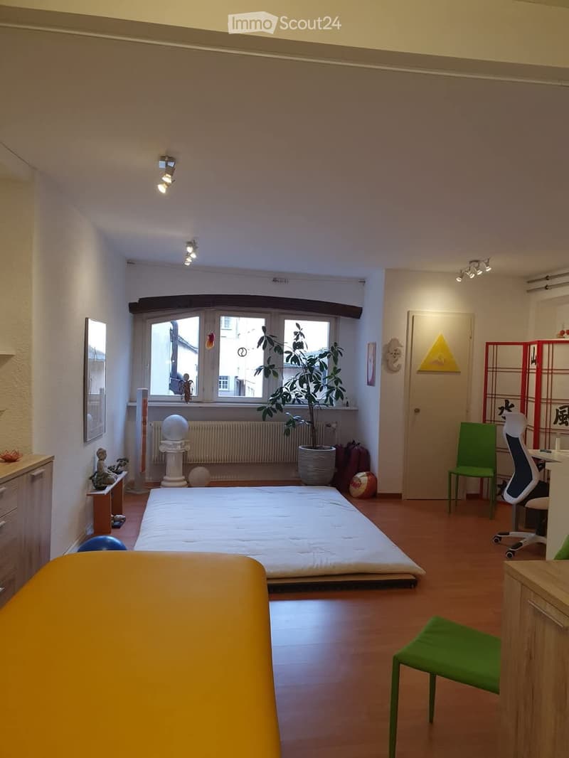 A well-lit bedroom with a tatami-style bed, a green chair, a desk with a chair, a potted plant, a mirror, and a small cabinet.