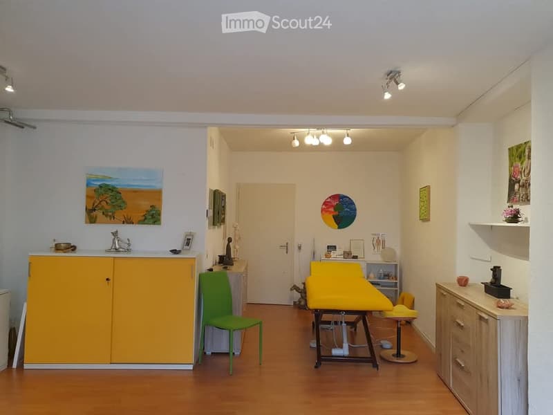 A well-lit room with a yellow cabinet, a green chair, a yellow examination table, a small stool, a sink with a faucet, shelves, and framed artwork on the walls.