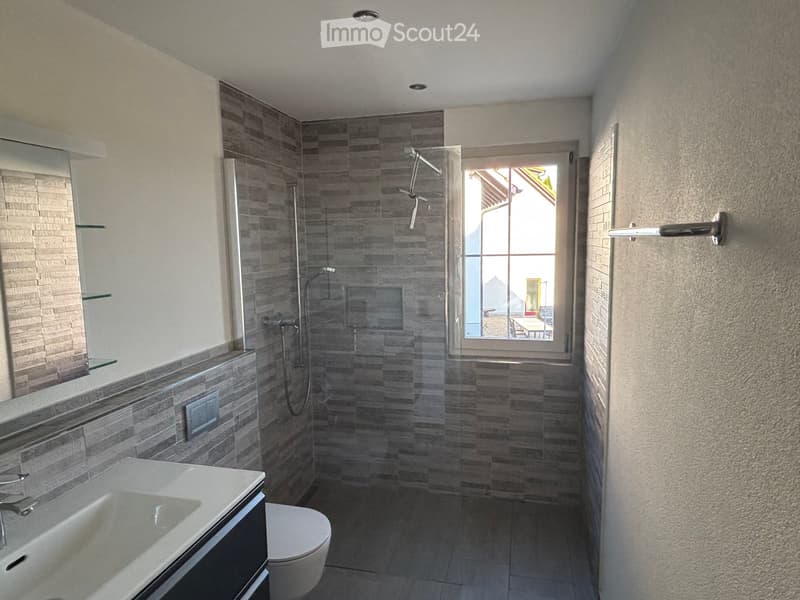 Modern bathroom with a white sink, toilet, large mirror, and a glass-enclosed shower with grey tiles. A window provides natural light.