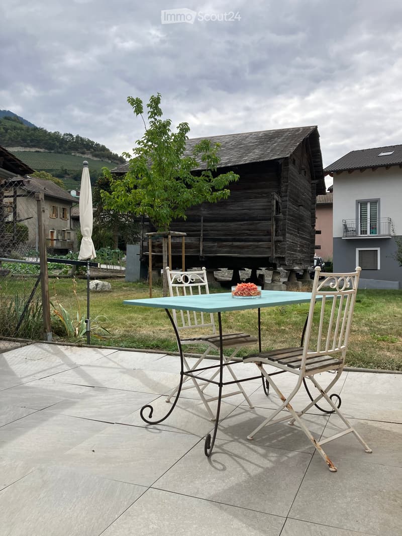 Outdoor patio with a light blue table, two white chairs, and a vase with pink flowers, surrounded by a grassy area and wooden structures in the background.