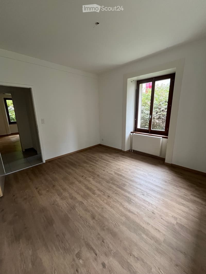 Empty room with wooden flooring, white walls, and a window with a radiator below it. The room has a door leading to another space.