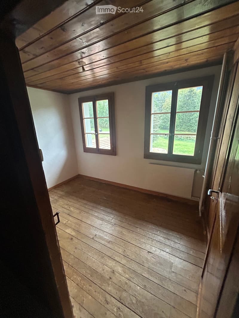 Empty room with wooden floor, wooden ceiling, and two windows showing greenery outside.