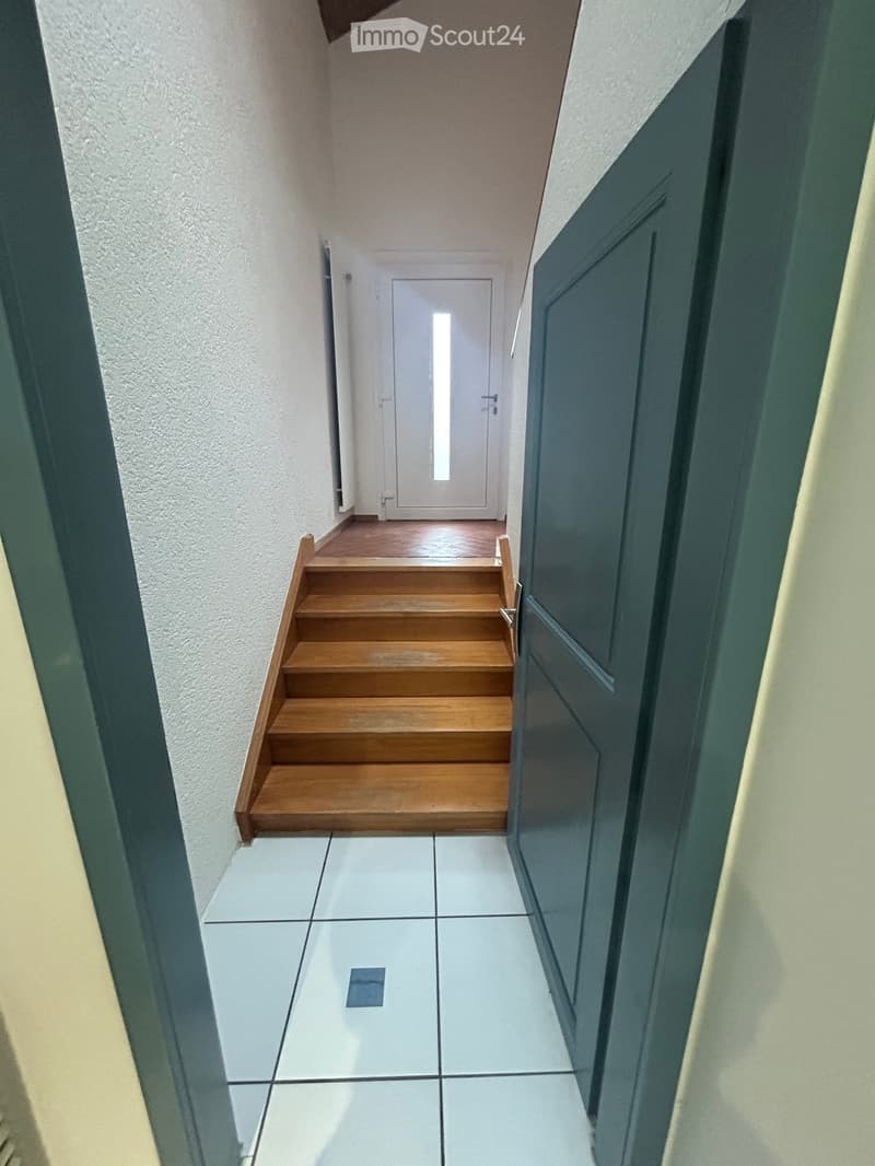 A narrow hallway with tiled floor and wooden staircase leading upwards, featuring white walls and a closed door at the end.