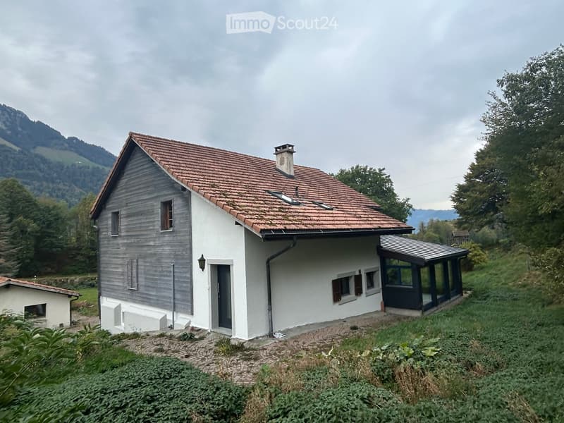 A small, single-story house with a red-tiled roof, white and grey exterior walls, and a chimney. The house features multiple windows, a front door, and a covered porch area with large glass panels. The surrounding area includes greenery, a paved pathway,