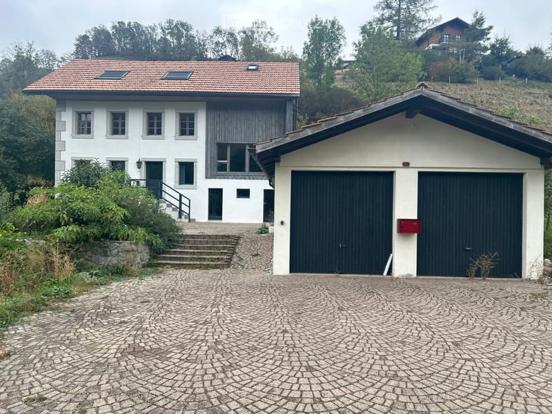 Two-story house with white exterior, red-tiled roof, and attached garage with black doors. The house features multiple windows and a small staircase leading to the entrance.