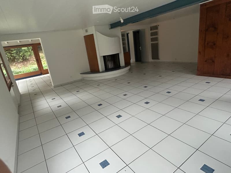 Empty room with white tiled floor and walls, featuring a fireplace and large windows with wooden frames.