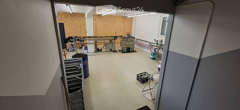 Empty industrial-style room with exposed brick wall, concrete floor, and various equipment and tools visible in the background.