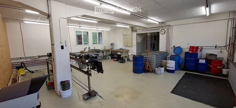 The image shows an empty industrial or workshop space with concrete floors, white walls, and large windows. The room contains various storage items such as barrels, crates, and tools, but no furniture or major fixtures are present.