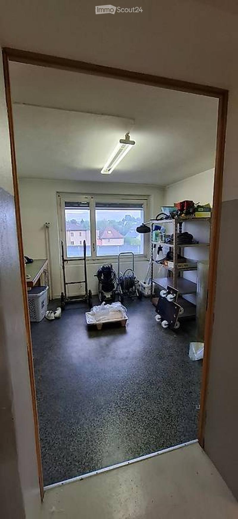 Empty storage room with concrete floor, white walls, large window, shelves, and various stored items including a stroller, boxes, and tools.