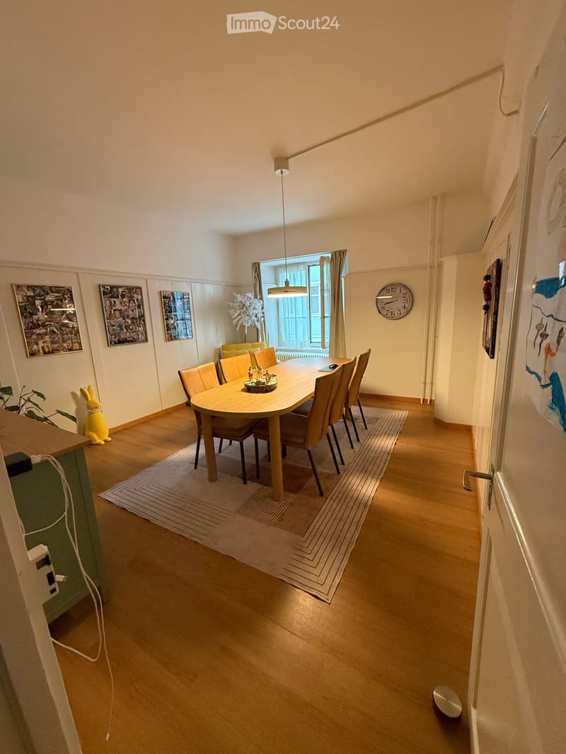 Dining room with a round wooden table surrounded by six chairs, a clock on the wall, picture frames, a small plant, and a toy rabbit on the floor.