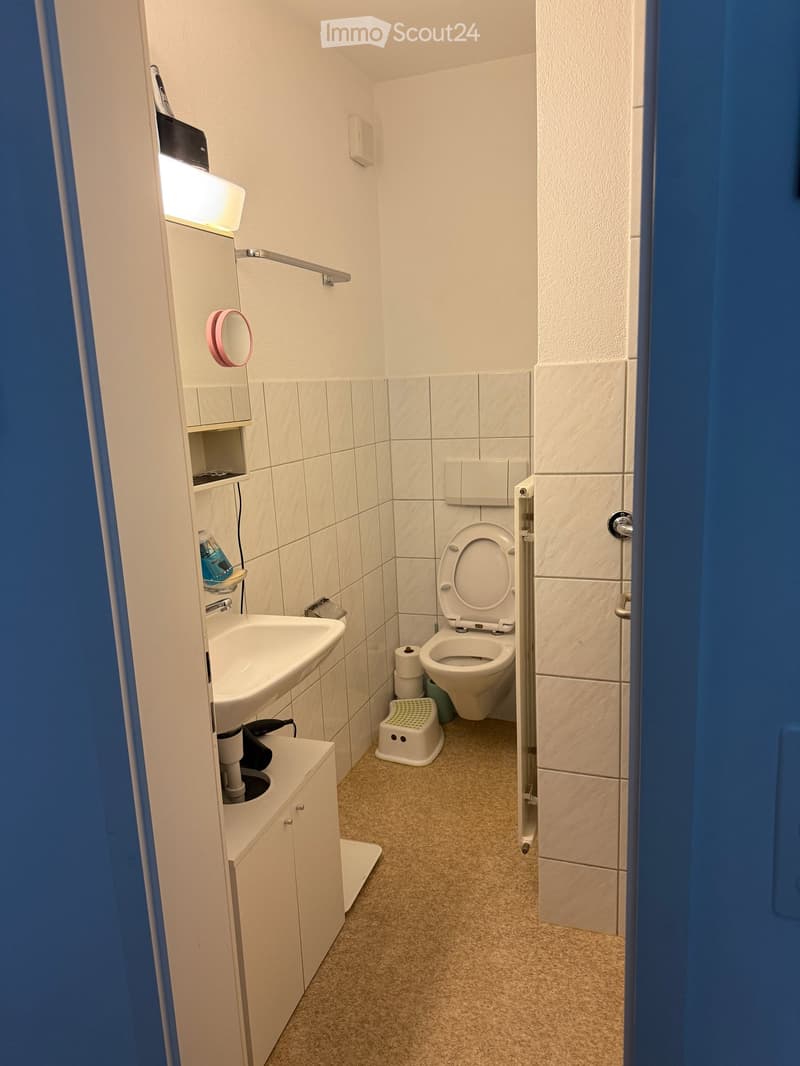 Compact bathroom with white tiled walls, a sink with a cabinet, a toilet, a mirror, and a small stool.