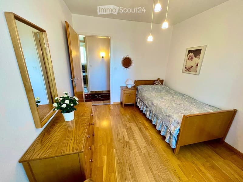 Cozy bedroom with wooden flooring, a single bed with patterned bedding, a wooden nightstand with a lamp, a dresser with a mirror, and a small flower pot on the dresser.