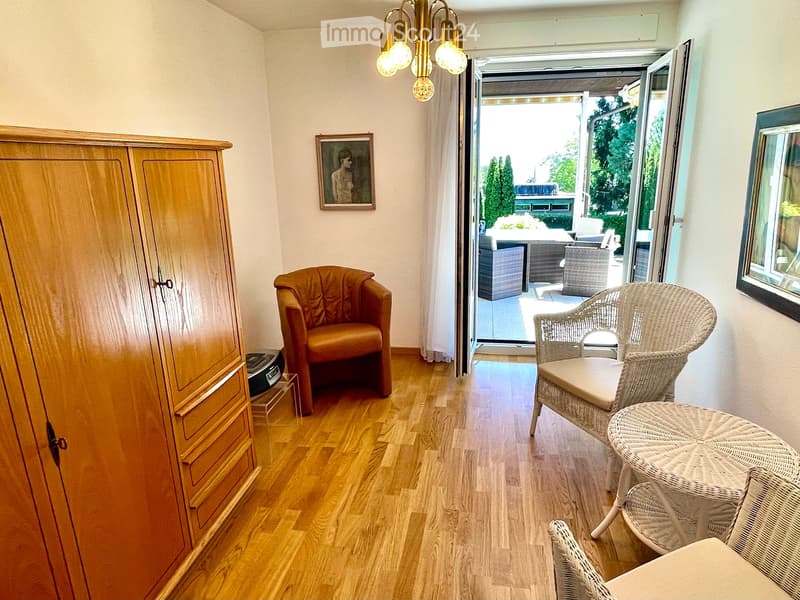 A furnished room with wooden flooring, a large wardrobe, an armchair, a small table, and a wicker chair near the balcony entrance.