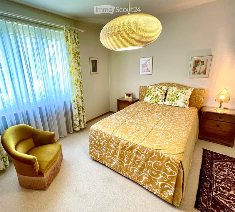 Cozy bedroom with a large bed, patterned bedspread, nightstands, a yellow armchair, and framed artwork on the walls.